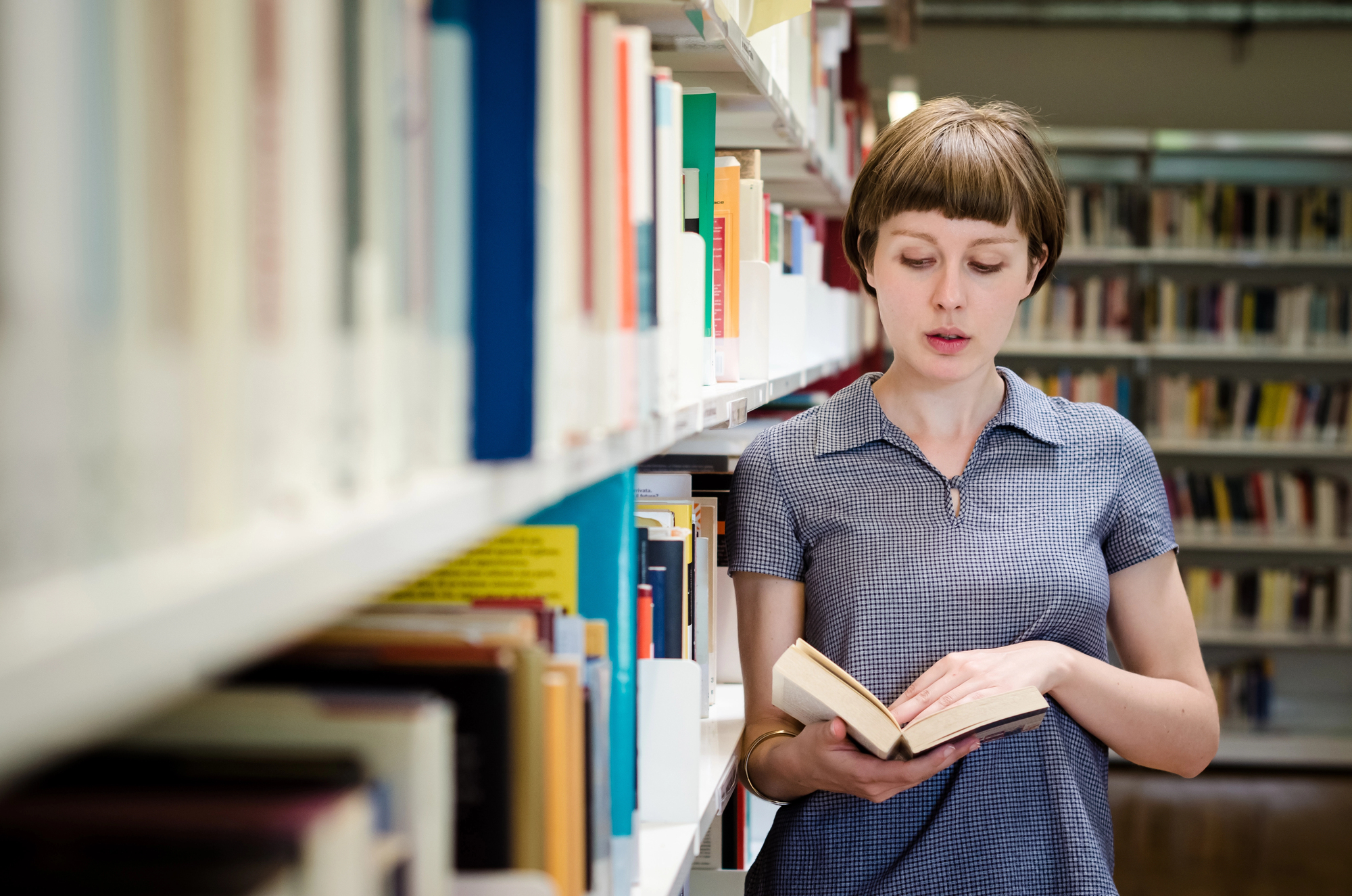 A person with short hair, wearing a blue collared shirt, stands in a library, reading a book. They are surrounded by tall shelves filled with books.