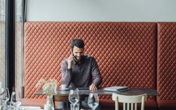 A man with a beard is sitting at a restaurant booth, talking on a smartphone. He is wearing a sweater and smiling, with a tablet and papers on the table. The booth has a quilted, orange-brown upholstered design, and the setting is bright and modern.