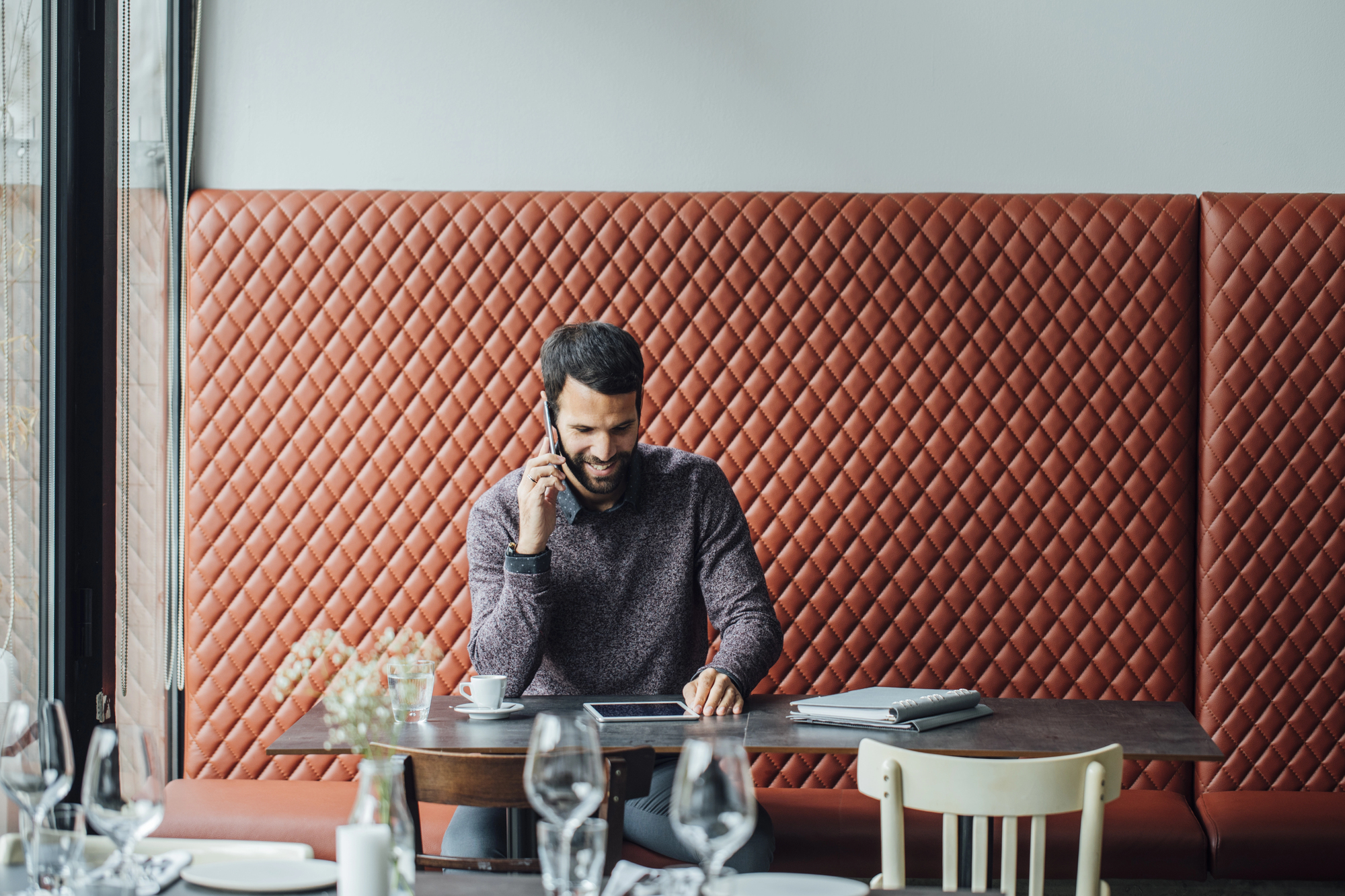 A man with a beard is sitting at a restaurant booth, talking on a smartphone. He is wearing a sweater and smiling, with a tablet and papers on the table. The booth has a quilted, orange-brown upholstered design, and the setting is bright and modern.