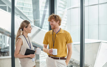 A man and woman having a conversation in a bright, modern office. Both are casually dressed, holding coffee mugs, and wearing lanyards. The woman holds a notebook. Large windows overlook a cityscape in the background.