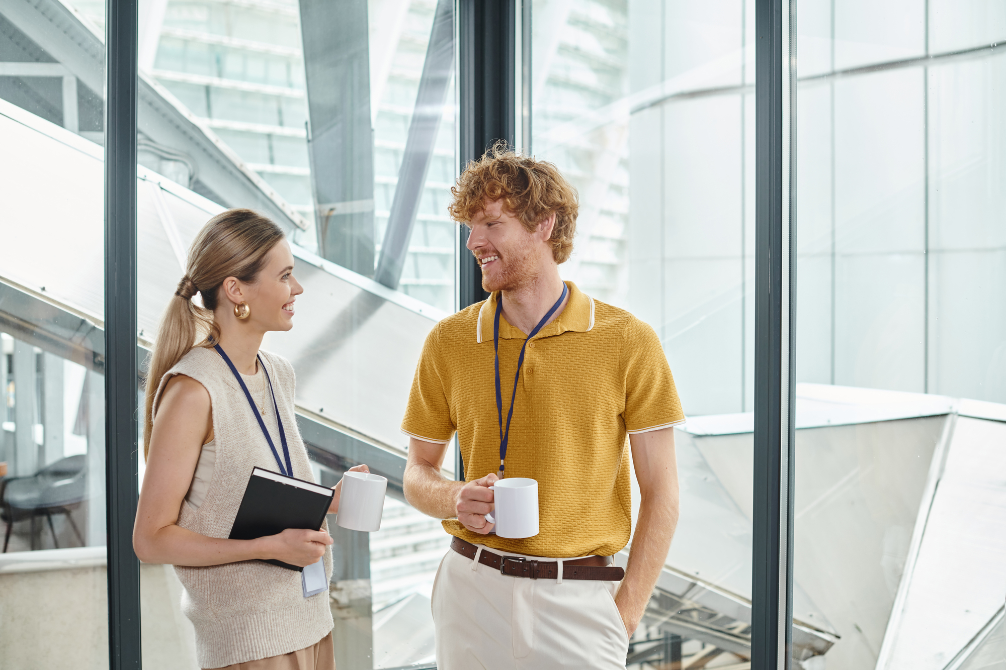 A man and woman having a conversation in a bright, modern office. Both are casually dressed, holding coffee mugs, and wearing lanyards. The woman holds a notebook. Large windows overlook a cityscape in the background.