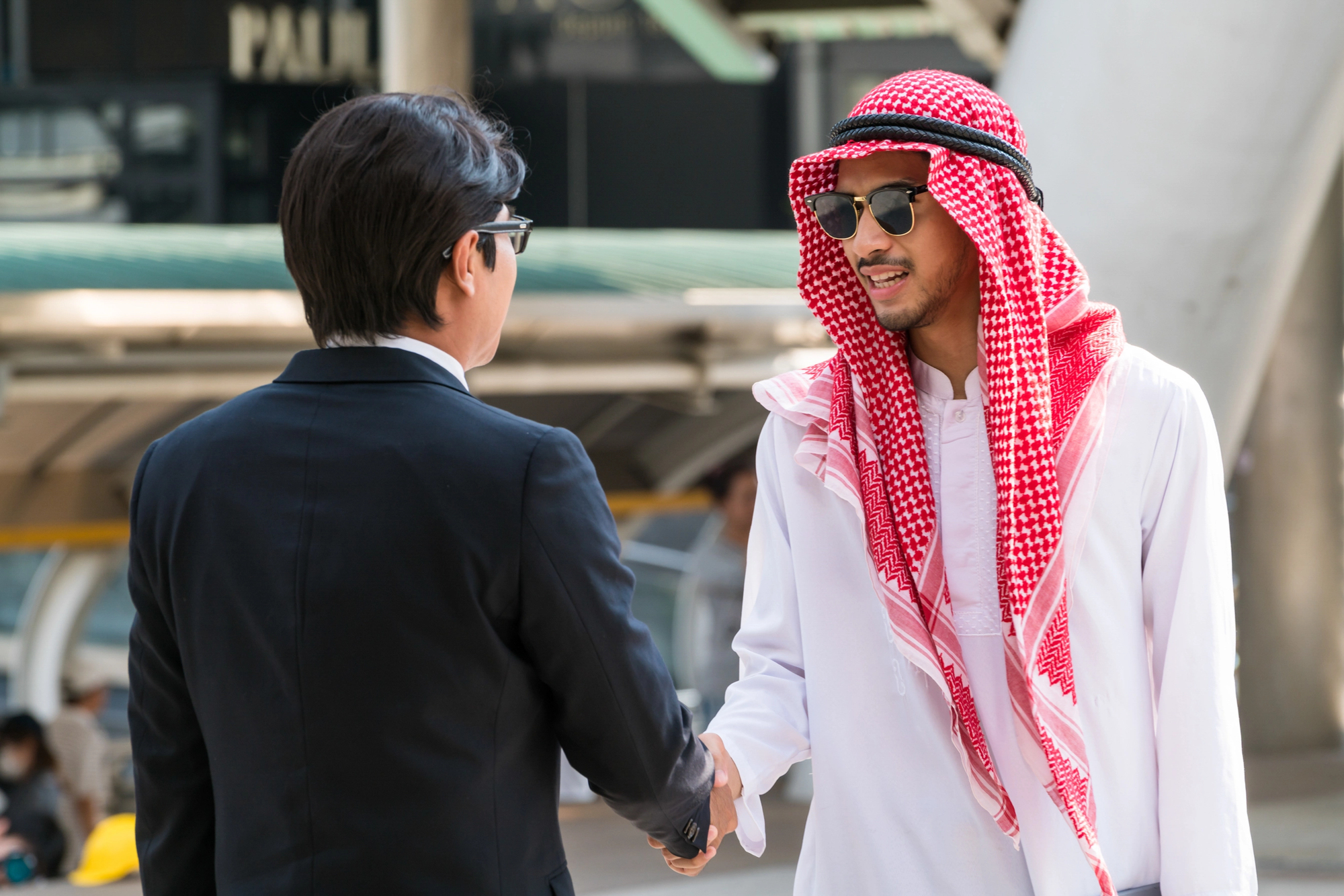 Two men shaking hands outdoors. One wears a suit and glasses, facing away from the camera. The other wears a red and white keffiyeh, a white thobe, and sunglasses. They are in a modern urban setting with blurred background elements.