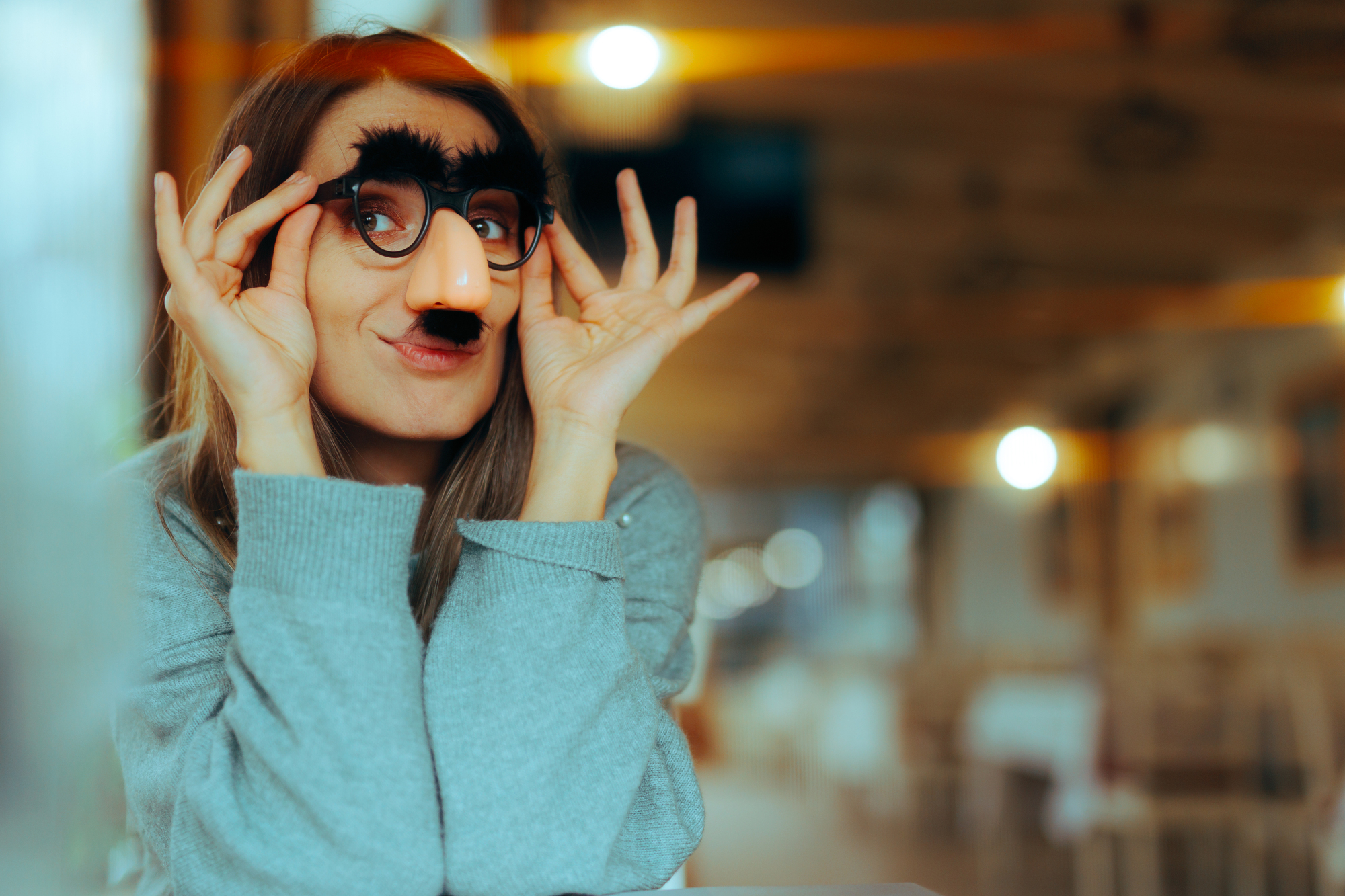 A person wearing a novelty disguise with thick black eyebrows, glasses, and a fake nose, smiles playfully while posing indoors. The background is softly blurred, suggesting a cozy café or restaurant setting.