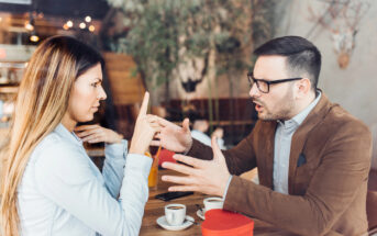 A woman and a man are sitting at a cafe table, engaged in a heated conversation. The woman gestures with her finger raised, while the man is speaking with his hands extended. Two cups of coffee and a red gift box are on the table.