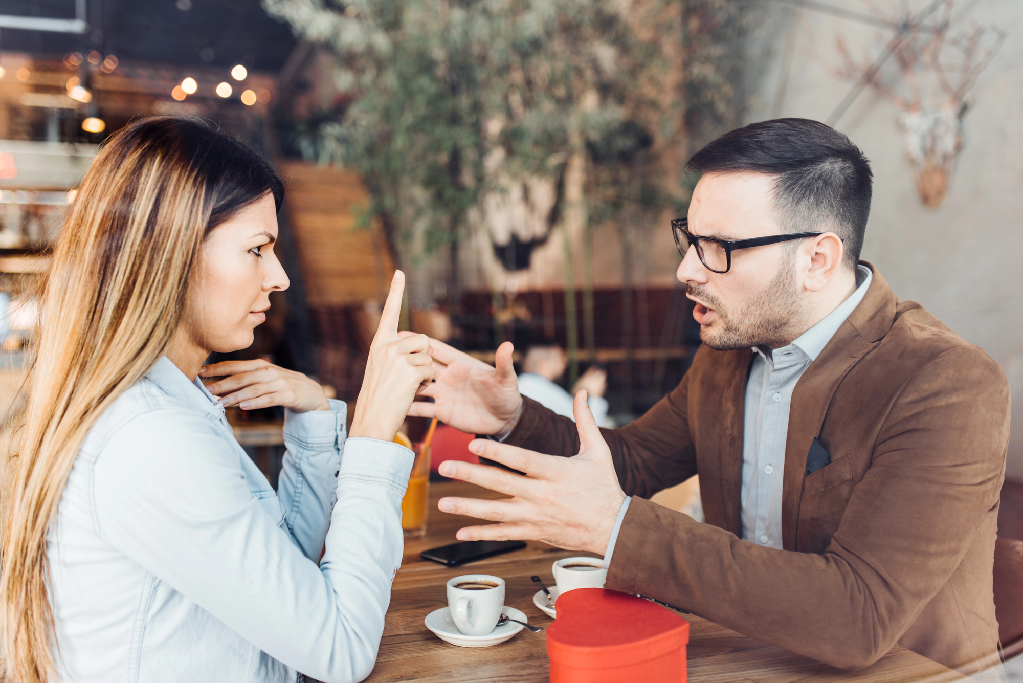 A woman and a man are sitting at a cafe table, engaged in a heated conversation. The woman gestures with her finger raised, while the man is speaking with his hands extended. Two cups of coffee and a red gift box are on the table.