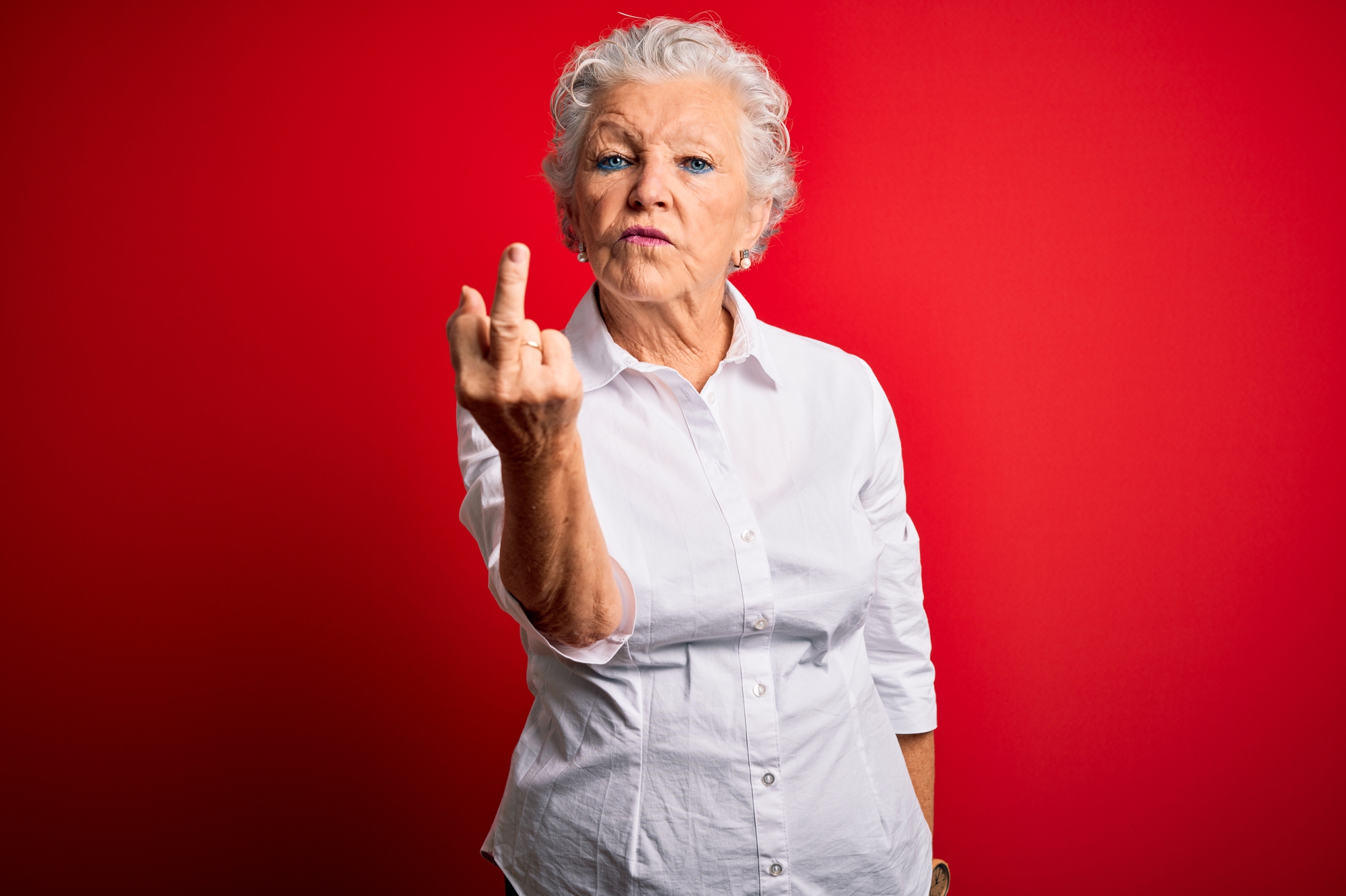 An older woman with white hair in a white shirt stands against a red background, making an assertive expression and gesturing with her middle finger raised.