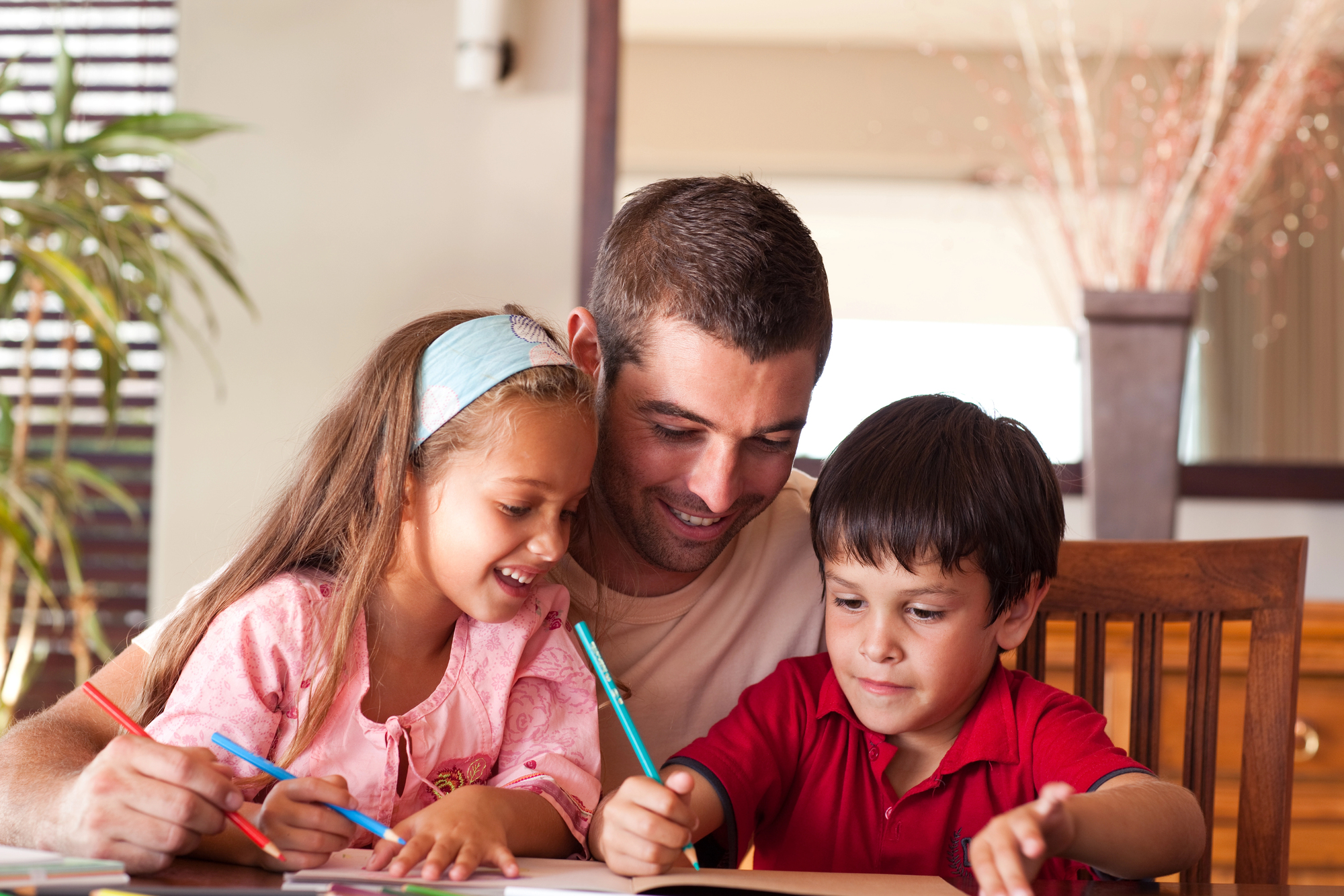 A man sits at a table with two children, a boy and a girl, who are drawing with colored pencils. They are smiling and appear to be enjoying the activity together in a cozy, well-lit room.