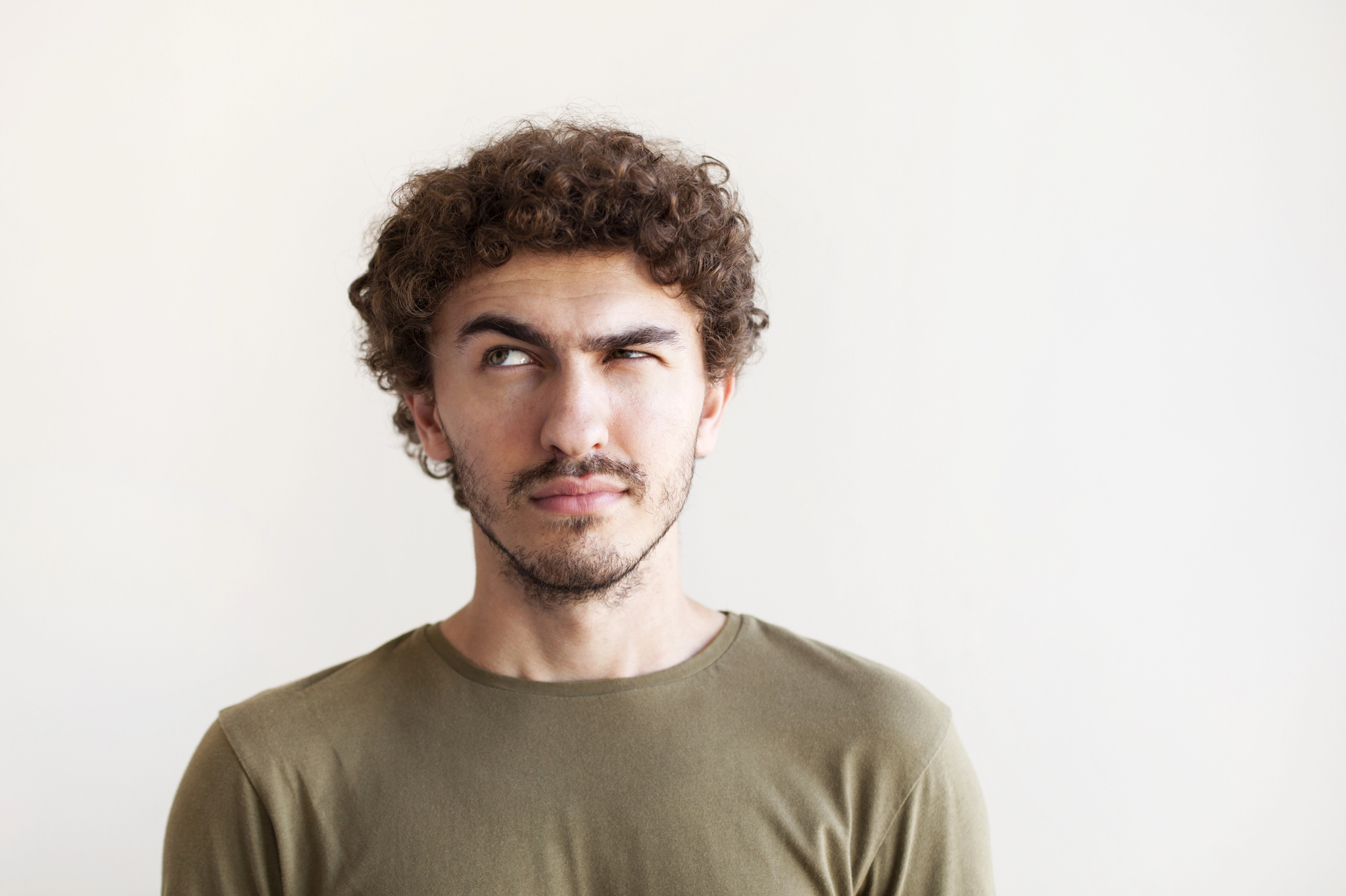 A young man with curly hair and a beard wearing a khaki shirt looks to the side against a neutral background.