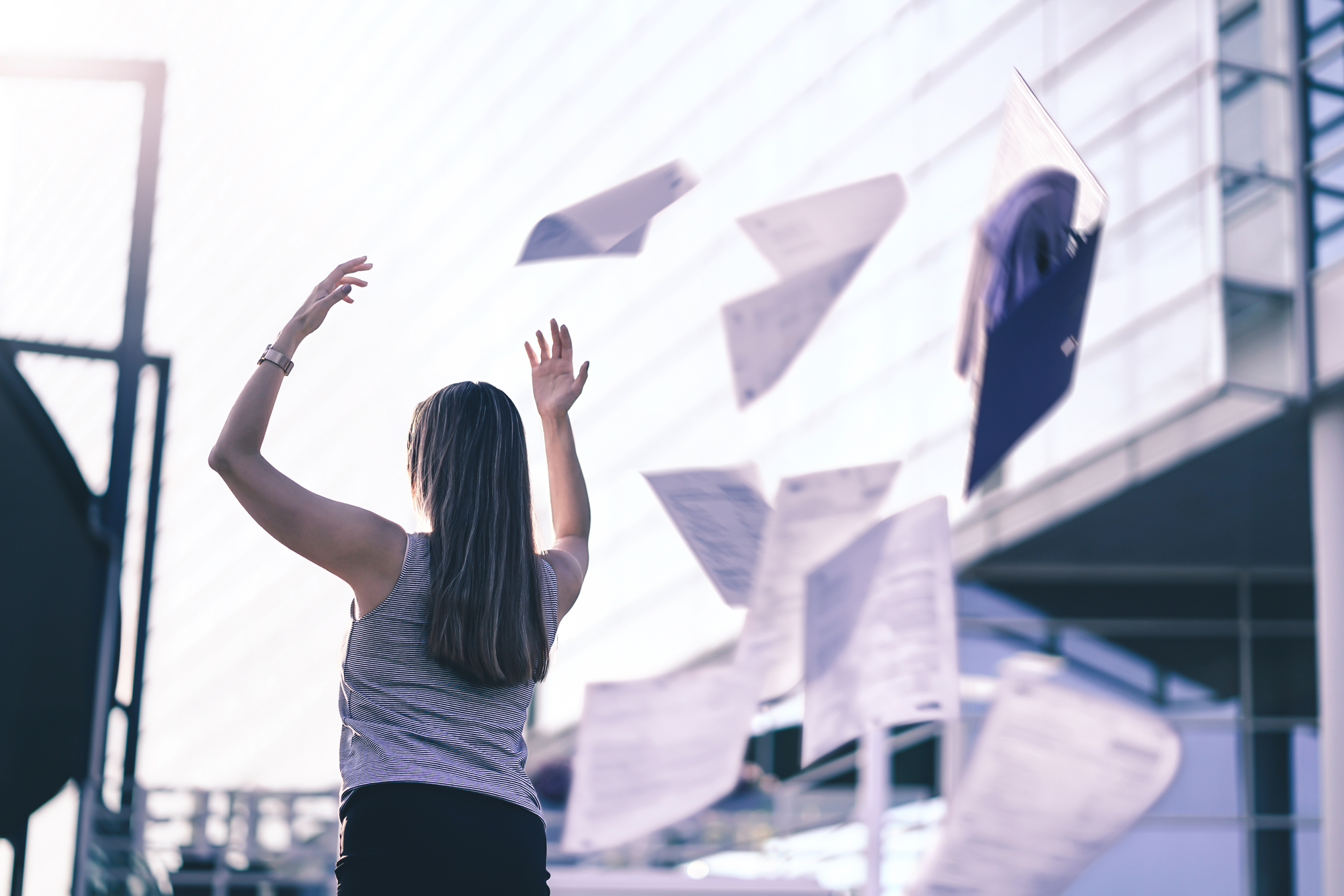 A person with long hair is standing outdoors, arms raised, while papers scatter in the air around them. The scene is set against a modern glass building, suggesting liberation or celebration.