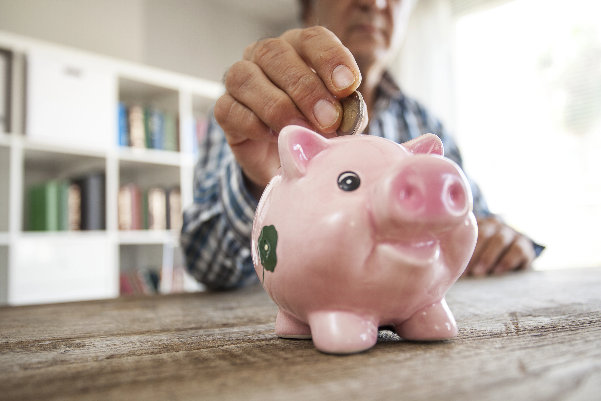 A person in a plaid shirt inserts a coin into a pink piggy bank on a wooden table. A bookshelf is visible in the background.