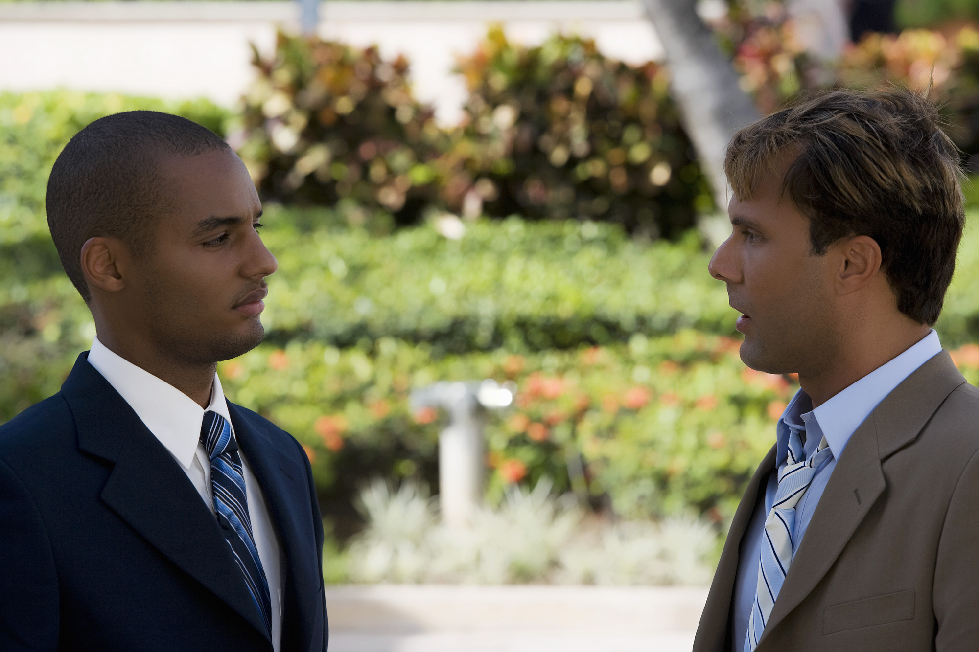 Two men in suits engaged in conversation outside, with a blurred background of greenery and flowers. Both appear focused on the discussion.