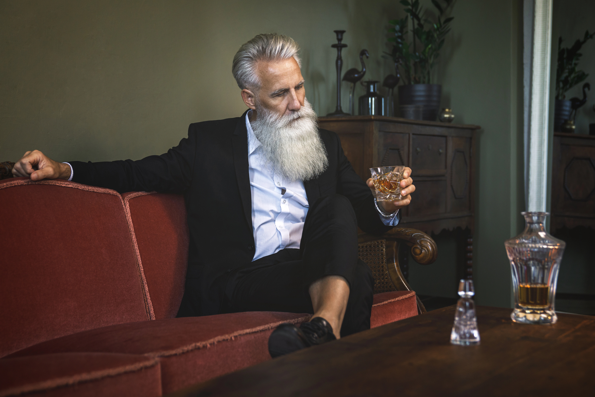 A man with a long white beard sits on a red sofa, holding a glass of whiskey. He wears a black suit with a white shirt. A decanter and an empty glass are on the wooden table in front of him. The room has dark, antique furniture and plants.