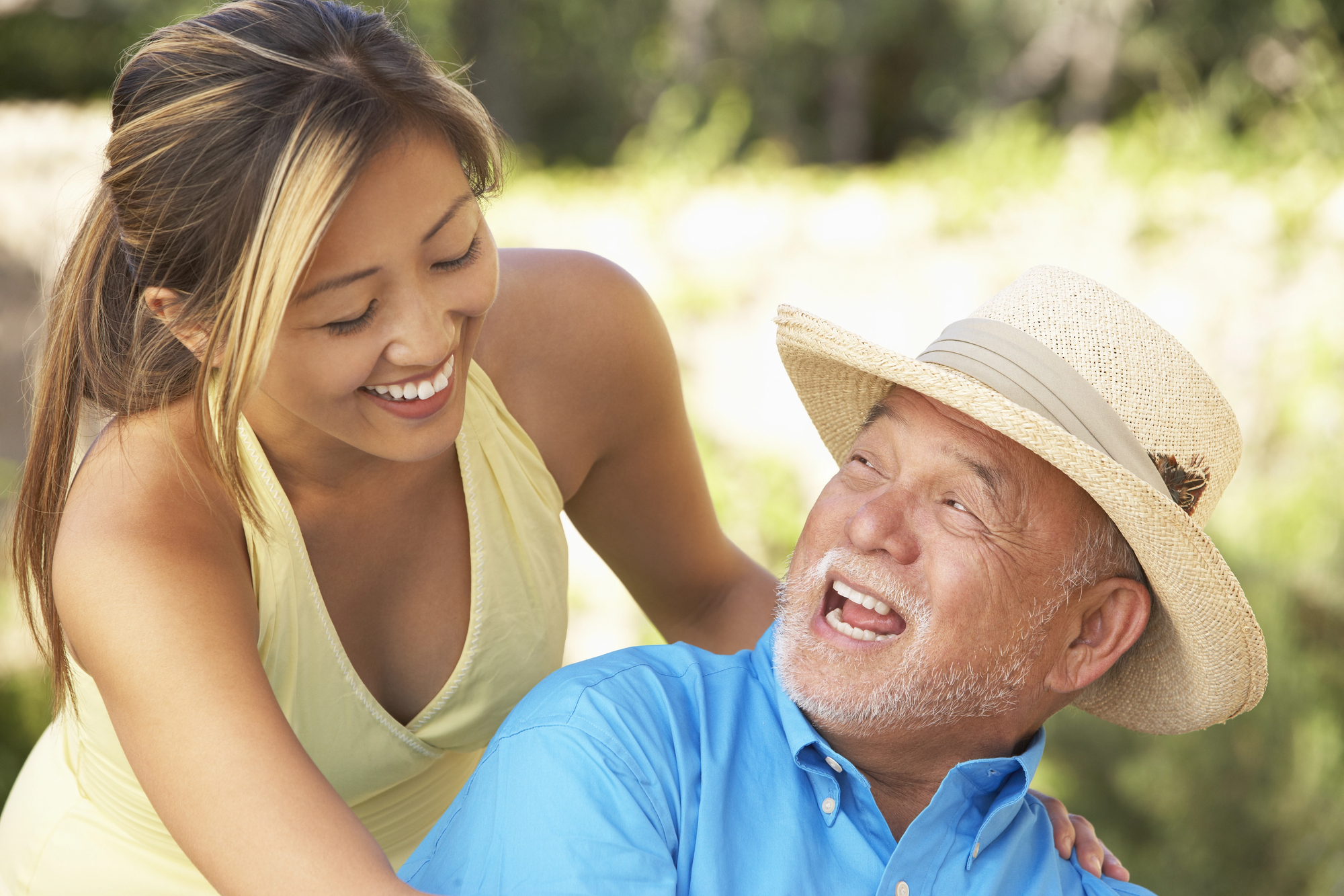 A smiling woman in a yellow tank top stands beside an older man wearing a straw hat and blue shirt. They are outside, with greenery in the background, sharing a joyful moment.