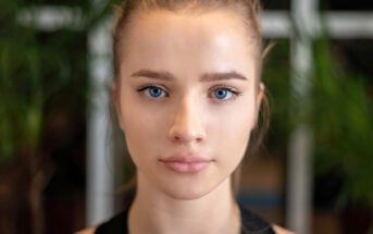 A woman with light hair pulled back looks directly at the camera. She has clear skin and blue eyes. The background is blurred with hints of greenery.