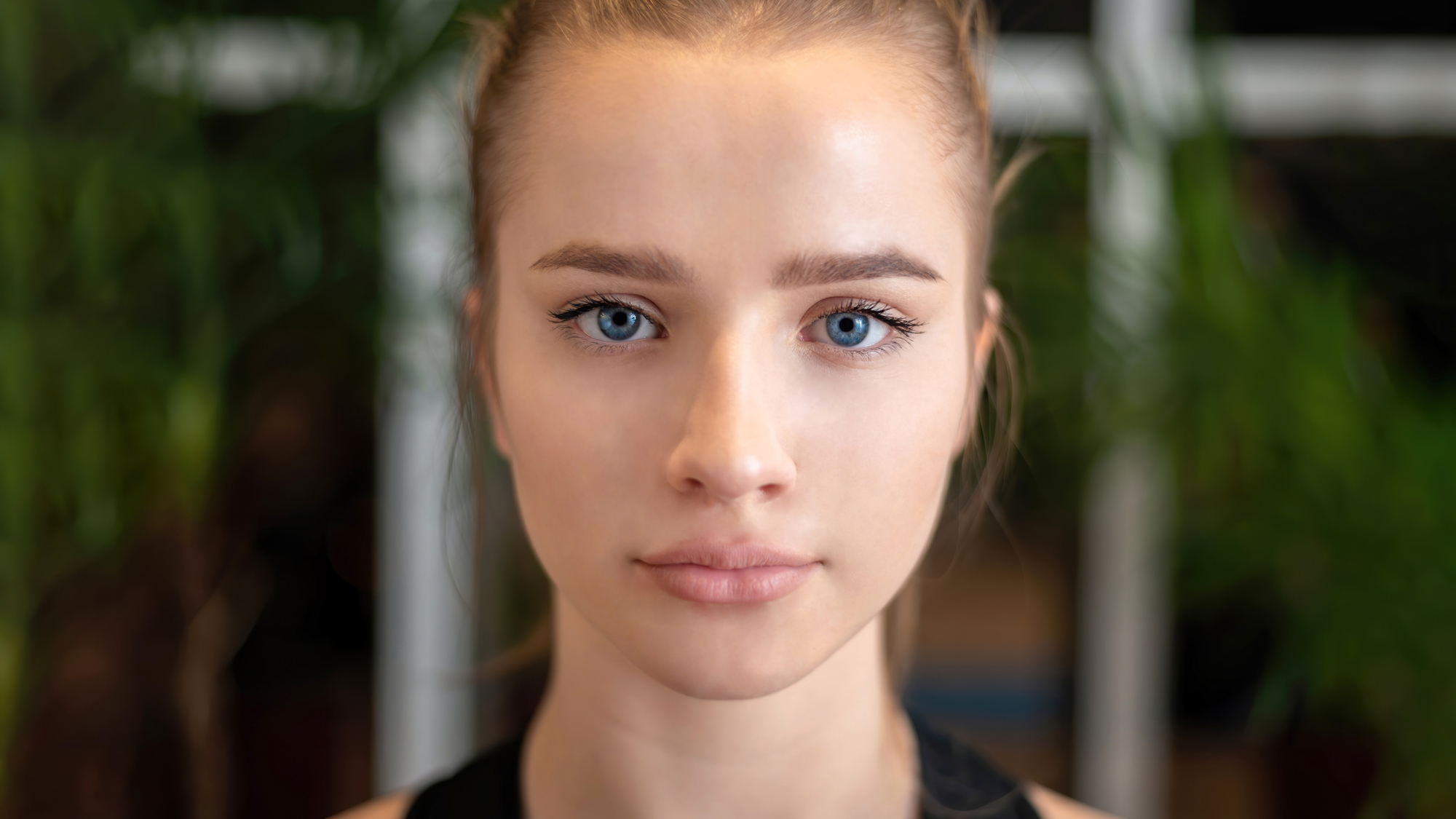 A woman with light hair pulled back looks directly at the camera. She has clear skin and blue eyes. The background is blurred with hints of greenery.