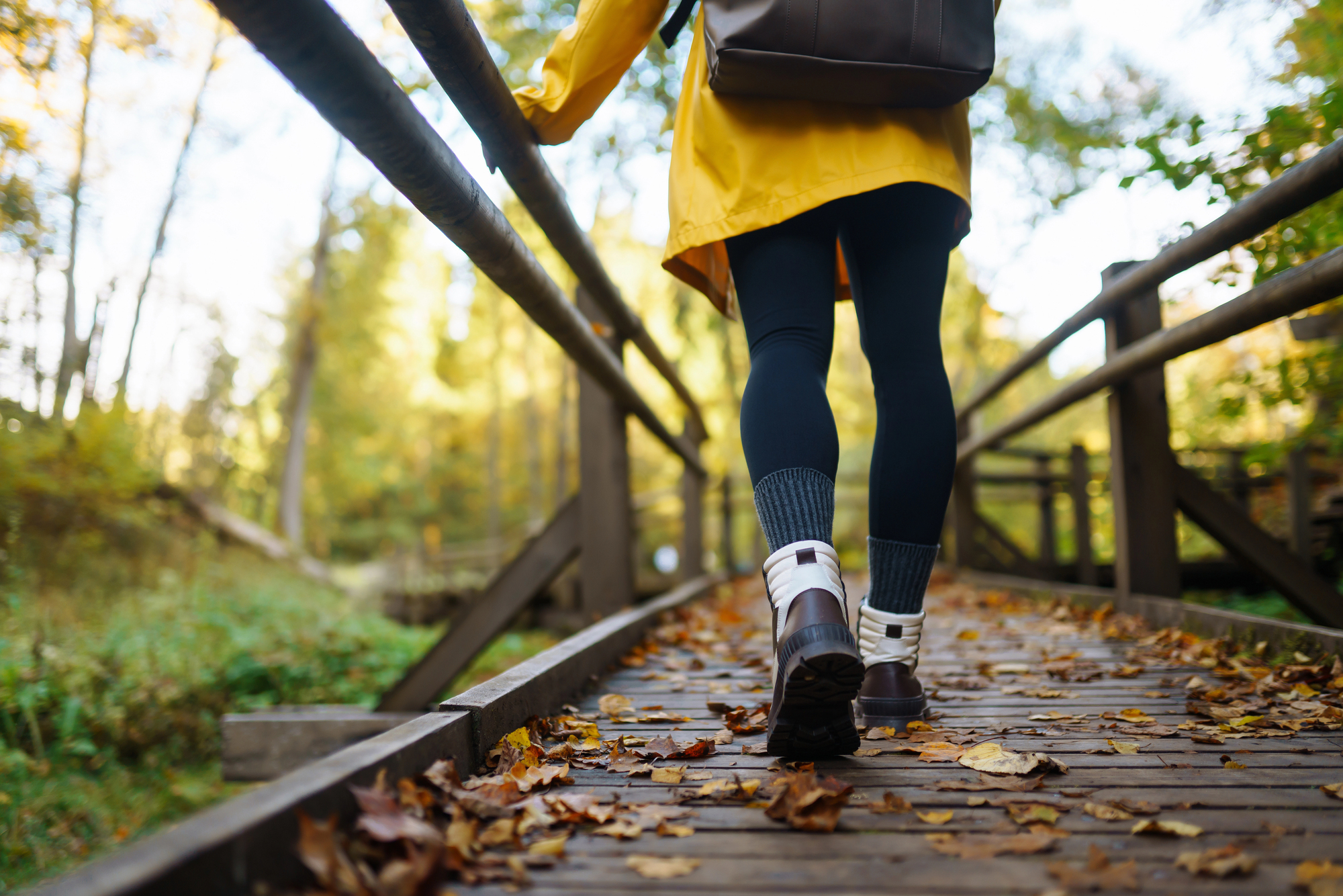 A person wearing a yellow jacket and warm boots walks across a wooden bridge covered with fallen autumn leaves, surrounded by trees in a forest. The scene captures the essence of a peaceful fall day outdoors.