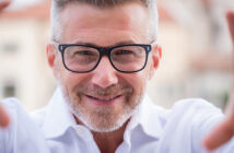 A smiling man with gray hair and a beard, wearing black glasses and a white shirt, appears to be holding a smartphone or camera to take a selfie. The background is softly blurred.