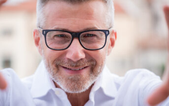 A smiling man with gray hair and a beard, wearing black glasses and a white shirt, appears to be holding a smartphone or camera to take a selfie. The background is softly blurred.