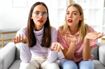 Two women are sitting on a couch with confused expressions. One has long dark hair and wears glasses and a light purple top, the other has blonde hair and wears a red and white striped shirt. Both have red lipstick and are gesturing with their hands.