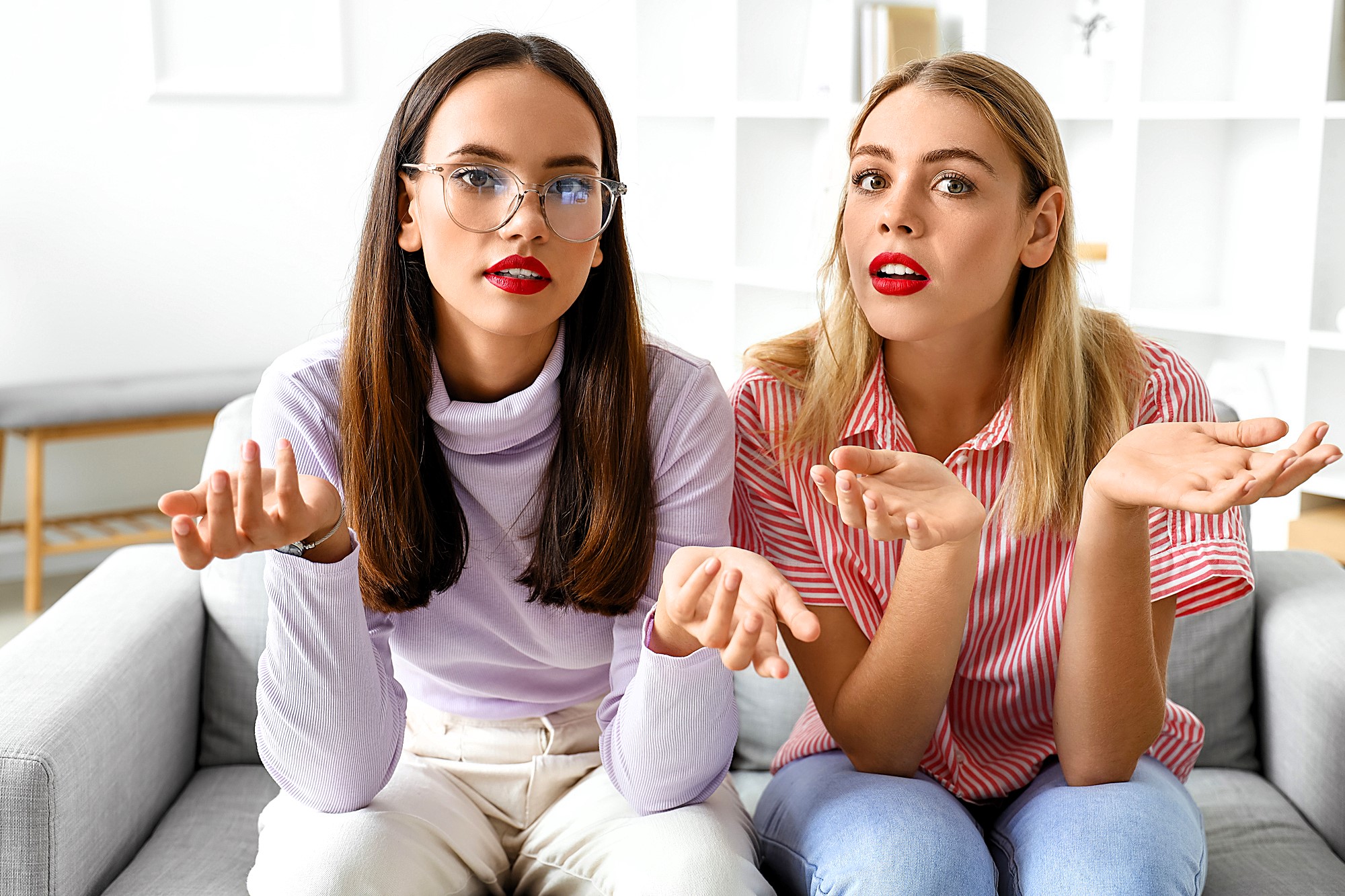 Two women are sitting on a couch with confused expressions. One has long dark hair and wears glasses and a light purple top, the other has blonde hair and wears a red and white striped shirt. Both have red lipstick and are gesturing with their hands.