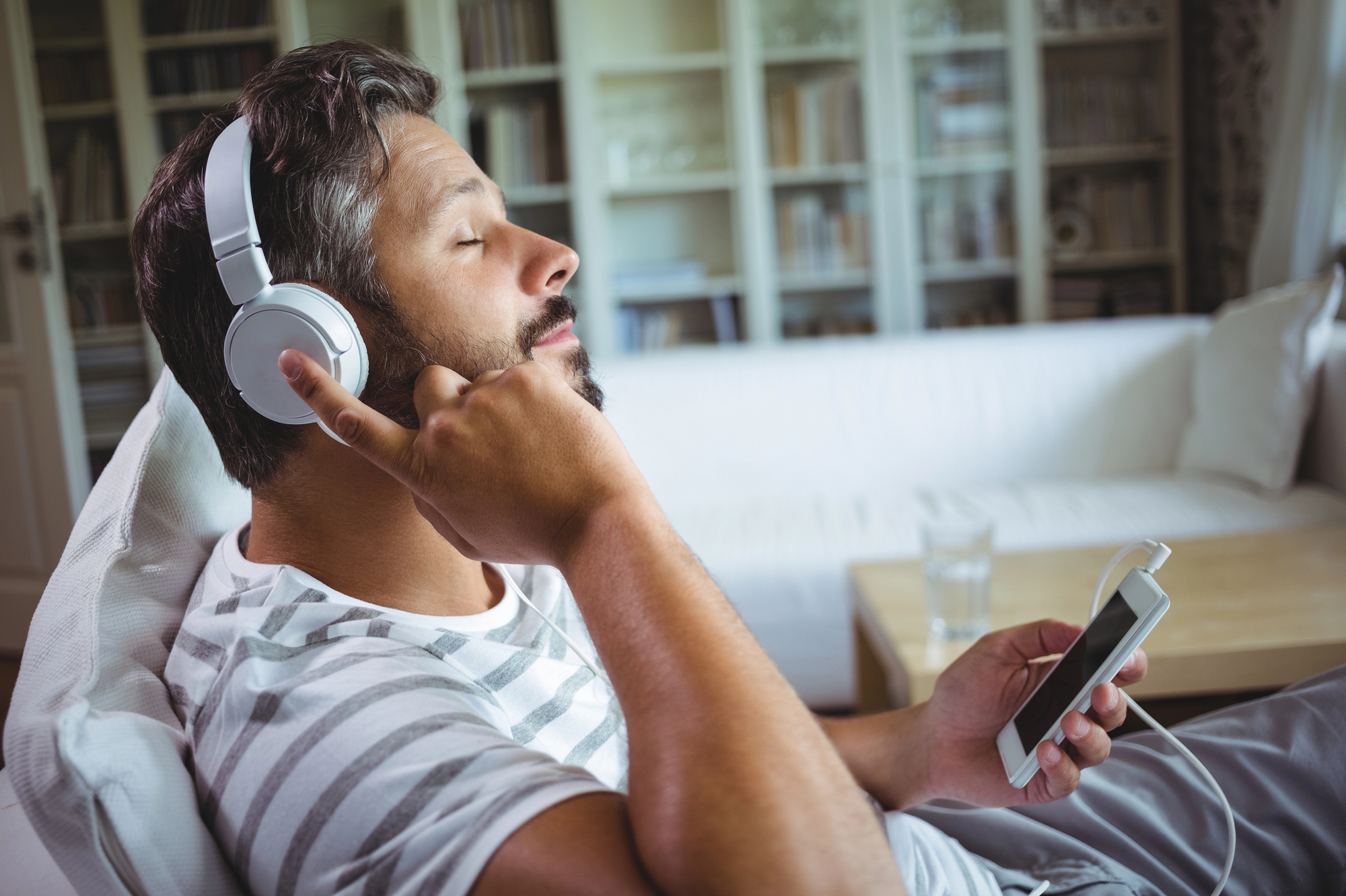 A man with headphones sits on a couch, eyes closed, enjoying music. He's holding a smartphone in one hand and appears relaxed in a bright living room with shelves in the background.