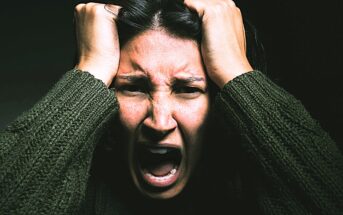 A person with long dark hair, wearing a dark textured sweater, is expressing intense emotion. They have their hands on their head and their mouth open, conveying a sense of frustration or anger. The background is dark.