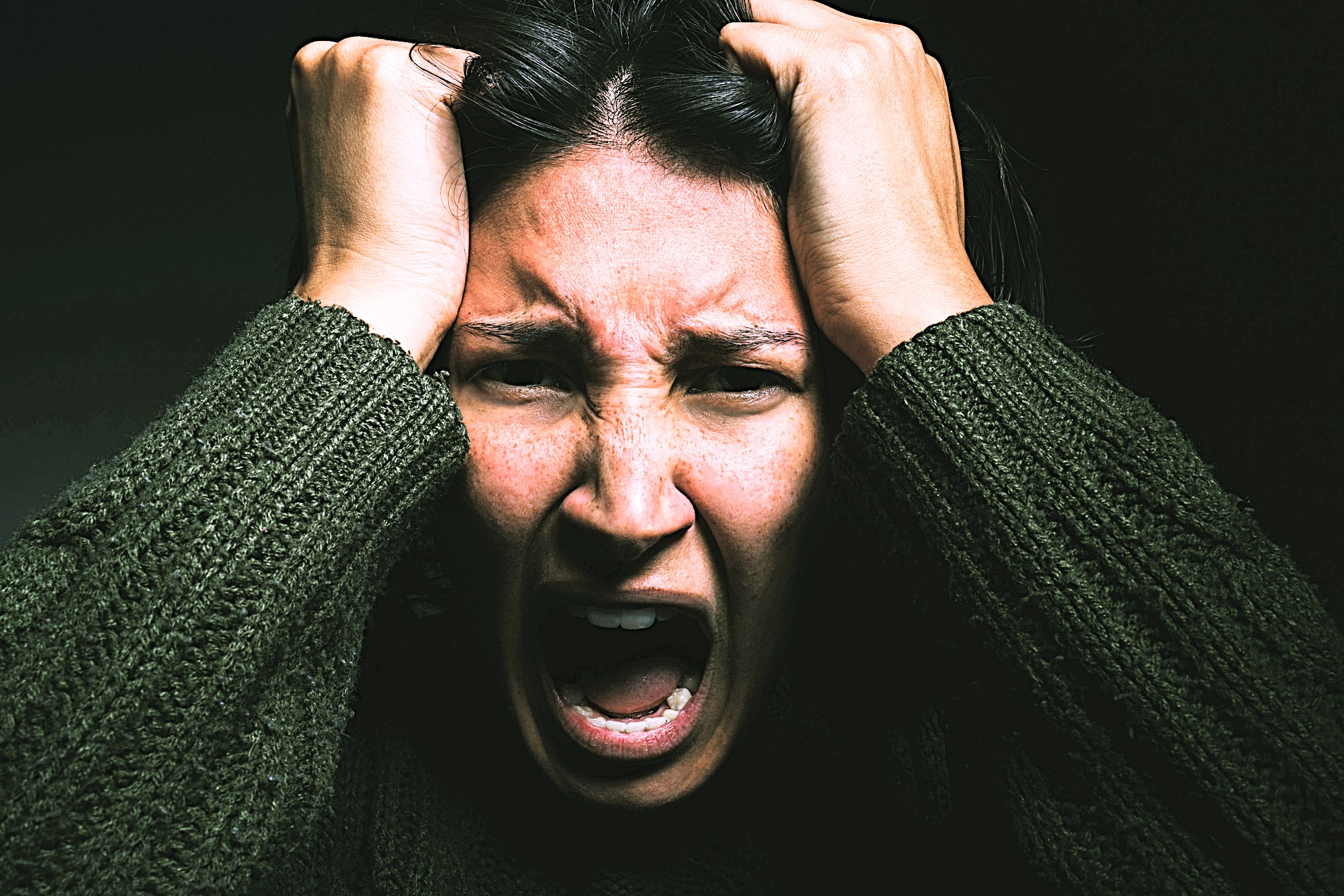 A person with long dark hair, wearing a dark textured sweater, is expressing intense emotion. They have their hands on their head and their mouth open, conveying a sense of frustration or anger. The background is dark.