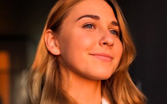 A person with long, light brown hair smiles softly in a warm, softly lit setting. They are wearing a small earring and a white top. The lighting casts a soft glow on their face.