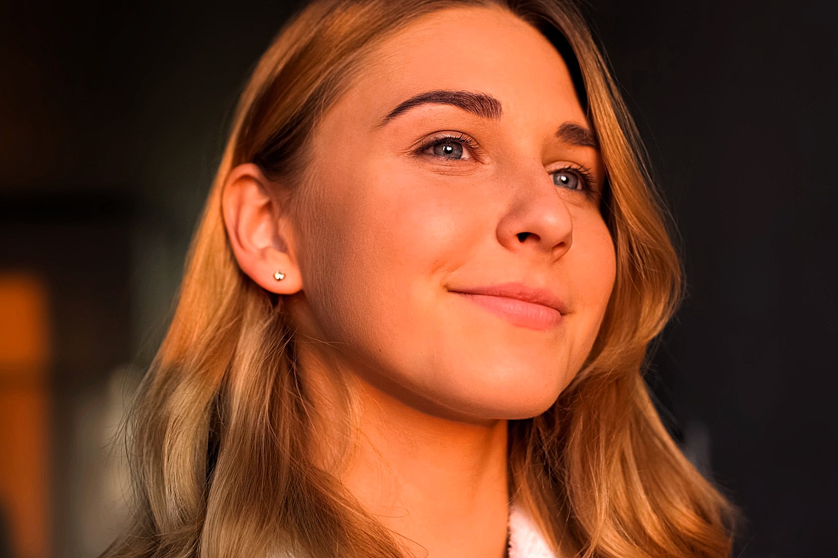 A person with long, light brown hair smiles softly in a warm, softly lit setting. They are wearing a small earring and a white top. The lighting casts a soft glow on their face.