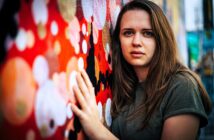 A person with long brown hair and wearing a green shirt leans against a vibrant, abstract mural with red, white, and black patterns. The background is slightly blurred, bringing focus to the person's thoughtful expression.