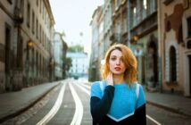 A woman with long hair stands on a quiet city street lined with traditional architecture. She is wearing a blue sweater and looking into the distance. The tram tracks and buildings create a symmetrical background.