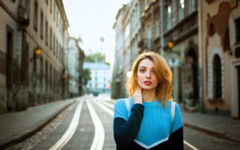 A woman with long hair stands on a quiet city street lined with traditional architecture. She is wearing a blue sweater and looking into the distance. The tram tracks and buildings create a symmetrical background.