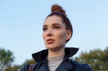 A woman with red hair is wearing a black leather jacket and looking into the distance. She stands outdoors with trees and a clear blue sky in the background.