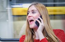 A woman with long blonde hair is smiling and talking on her smartphone. She is wearing a red sweater and standing in front of a window, with a blurred yellow background outside.