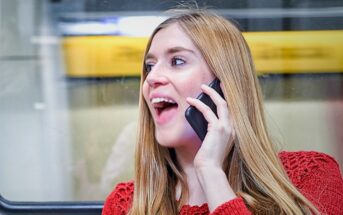 A woman with long blonde hair is smiling and talking on her smartphone. She is wearing a red sweater and standing in front of a window, with a blurred yellow background outside.