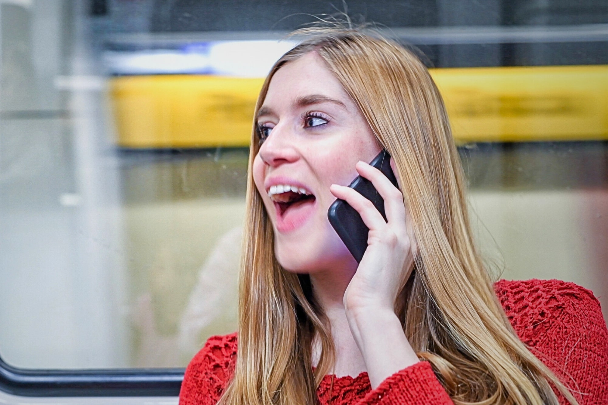 A woman with long blonde hair is smiling and talking on her smartphone. She is wearing a red sweater and standing in front of a window, with a blurred yellow background outside.