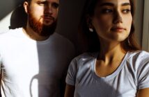 A man with a beard and a woman with long hair, both wearing white t-shirts, stand in partial sunlight. The woman looks out of frame while the man gazes downward. Shadows create dramatic lighting across the scene.