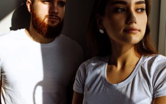 A man with a beard and a woman with long hair, both wearing white t-shirts, stand in partial sunlight. The woman looks out of frame while the man gazes downward. Shadows create dramatic lighting across the scene.