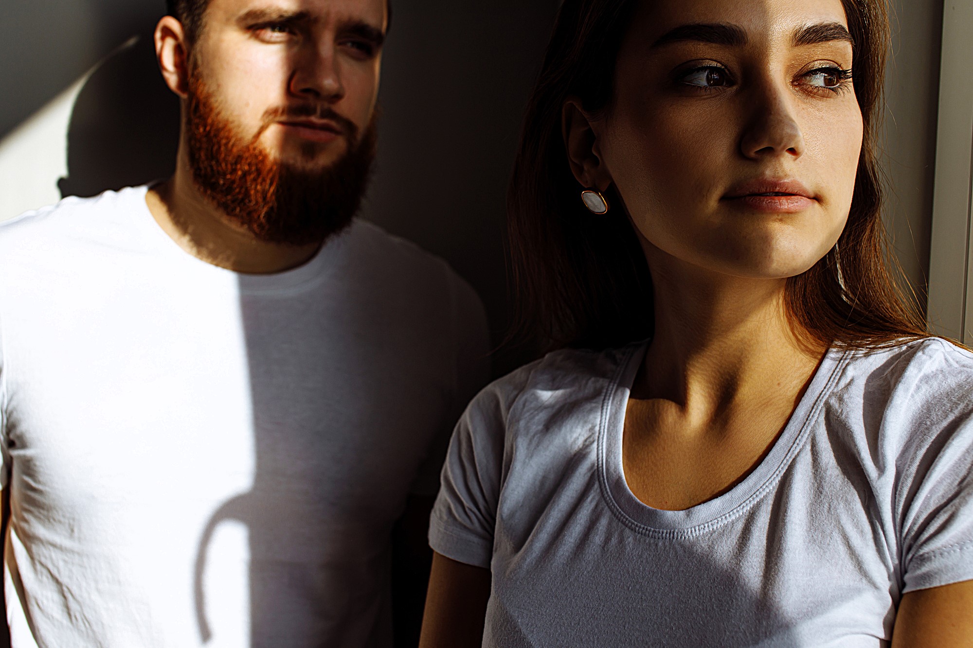 A man with a beard and a woman with long hair, both wearing white t-shirts, stand in partial sunlight. The woman looks out of frame while the man gazes downward. Shadows create dramatic lighting across the scene.