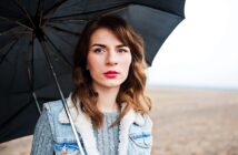 A person with long brown hair stands under a black umbrella in an open field. They are wearing a blue denim jacket with a fur collar and a gray sweater. The sky is overcast.