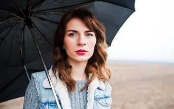 A person with long brown hair stands under a black umbrella in an open field. They are wearing a blue denim jacket with a fur collar and a gray sweater. The sky is overcast.