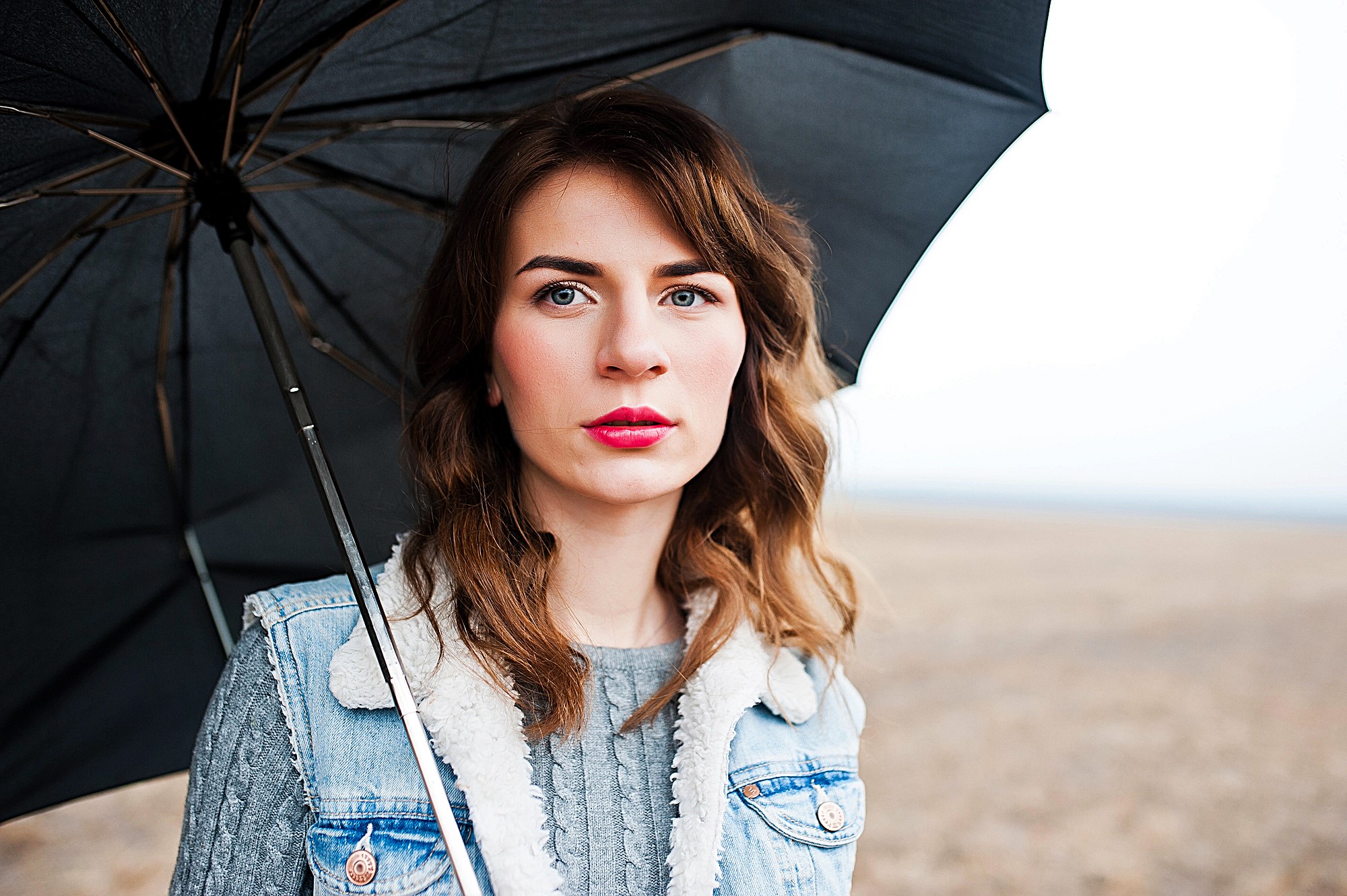 A person with long brown hair stands under a black umbrella in an open field. They are wearing a blue denim jacket with a fur collar and a gray sweater. The sky is overcast.