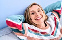 A person with long hair is smiling and leaning back with their hands behind their head. They are wearing a colorful striped sweater and sitting against a light blue background.