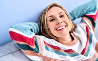 A person with long hair is smiling and leaning back with their hands behind their head. They are wearing a colorful striped sweater and sitting against a light blue background.