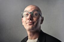 A bald man with glasses makes a quirky expression while wearing a black blazer and white shirt against a gray background.