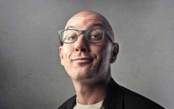 A bald man with glasses makes a quirky expression while wearing a black blazer and white shirt against a gray background.