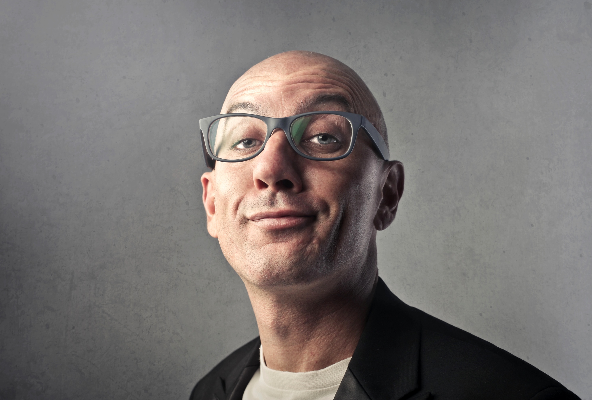 A bald man with glasses makes a quirky expression while wearing a black blazer and white shirt against a gray background.