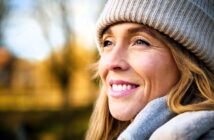 Close-up of a smiling woman wearing a knitted hat and scarf. She looks content and is gazing into the distance, with a blurred outdoor background and soft, natural lighting.