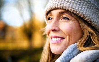 Close-up of a smiling woman wearing a knitted hat and scarf. She looks content and is gazing into the distance, with a blurred outdoor background and soft, natural lighting.