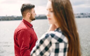 A man in a red shirt looks thoughtfully at a body of water while a woman in a checkered shirt stands in the foreground, slightly out of focus. The scene conveys a sense of contemplation near a waterfront.